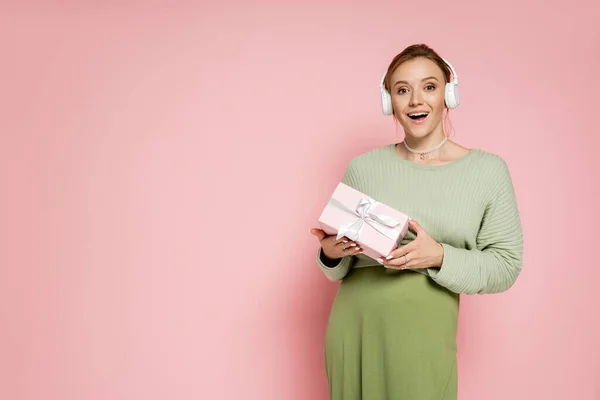 Mujer embarazada con estilo en auriculares con caja de regalo sobre fondo rosa - foto de stock