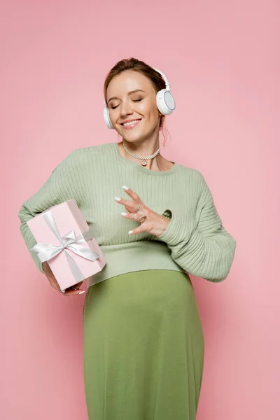 Trendige schwangere Frau mit Kopfhörer hält Geschenk isoliert auf rosa — Stockfoto