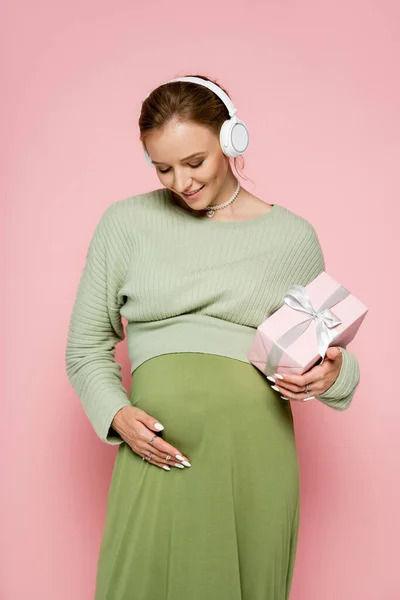 Mulher grávida elegante ouvindo música em fones de ouvido e segurando presente no fundo rosa — Fotografia de Stock
