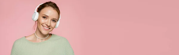 Позитивна жінка в навушниках і зелений светр дивиться на камеру ізольовано на рожевому, банер — стокове фото