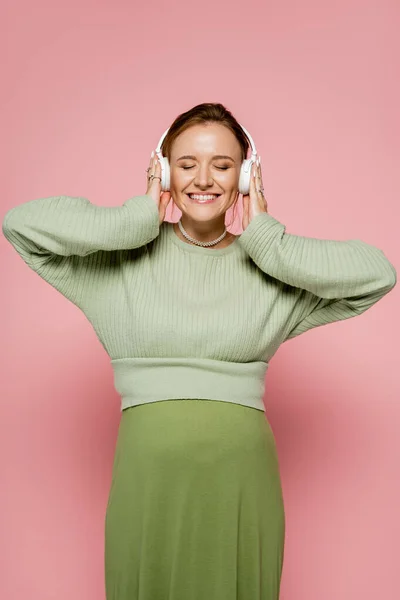 Веселая беременная женщина в свитере слушает музыку в наушниках на розовом фоне — стоковое фото