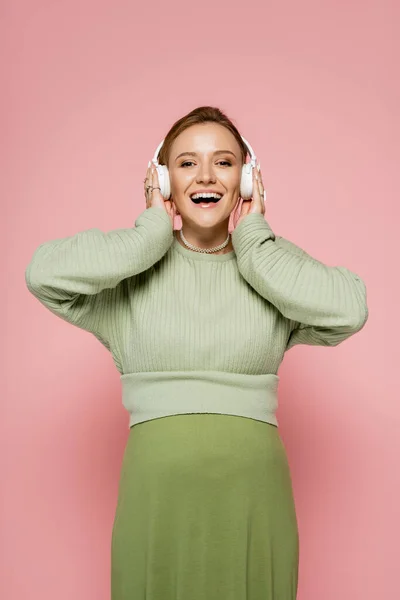 Захоплена вагітна жінка слухає музику в навушниках і дивиться на камеру на рожевому фоні — стокове фото