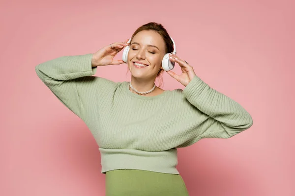 Mulher grávida positiva fechando os olhos enquanto ouve música em fones de ouvido no fundo rosa — Fotografia de Stock