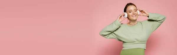 Веселая беременная женщина в зеленом наряде слушает музыку в наушниках на розовом фоне, баннер — стоковое фото