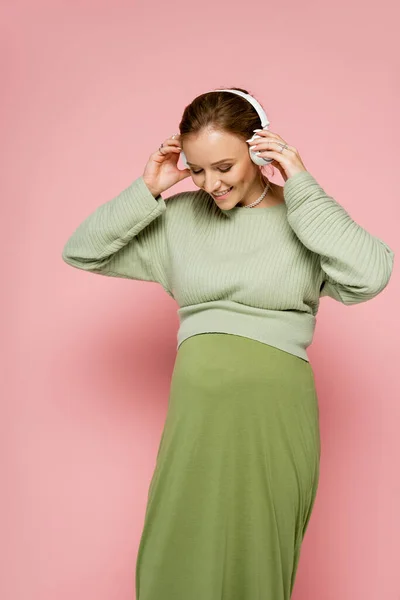 Mujer embarazada feliz en suéter verde con auriculares y mirando el vientre sobre fondo rosa - foto de stock