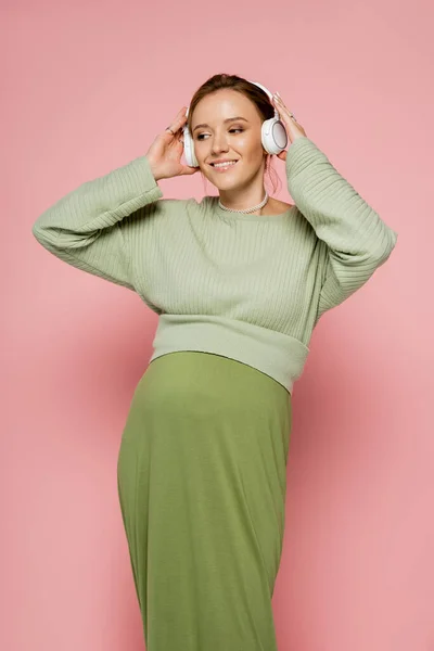 Щаслива вагітна жінка в навушниках дивиться геть, слухаючи музику на рожевому фоні — стокове фото