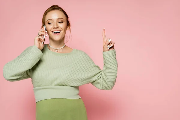 Веселая беременная женщина в свитере разговаривает по смартфону и указывает пальцем на розовый фон — стоковое фото