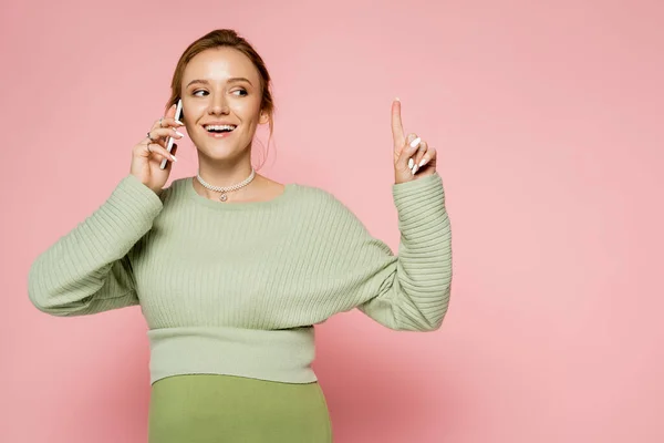 Улыбающаяся беременная женщина в свитере разговаривает по мобильному телефону и показывает пальцем на розовом фоне — стоковое фото