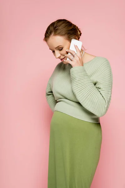 Mujer bastante embarazada en traje verde hablando en teléfono inteligente aislado en rosa - foto de stock