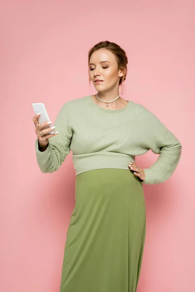 Young pregnant woman in green outfit using smartphone and holding hand on hip on pink background — Stock Photo