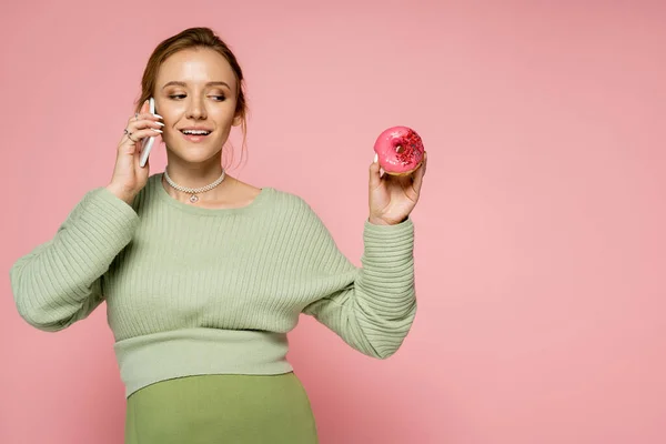 Молодая беременная женщина разговаривает на смартфоне и глядя на пончик изолирован на розовый — стоковое фото