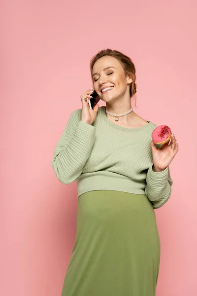 Веселая беременная женщина разговаривает на смартфоне и держит пончик на розовом фоне — стоковое фото