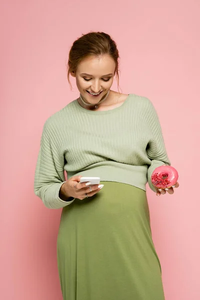 Pregnant woman in green outfit using smartphone and holding donut isolated on pink — Stock Photo