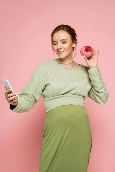Беременная женщина делает селфи и держит вкусный пончик на розовом фоне — стоковое фото