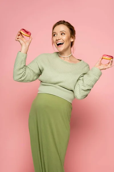 Возбужденная беременная женщина в свитере держит сладкие пончики на розовом фоне — стоковое фото