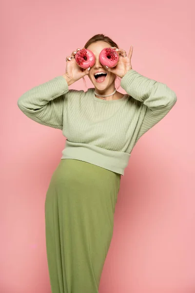 Mujer embarazada emocionada cubriendo la cara con rosquillas sobre fondo rosa - foto de stock