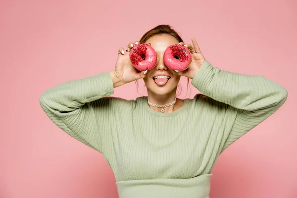 Jovem mulher saindo da língua e segurando donuts perto de olhos isolados em rosa — Fotografia de Stock