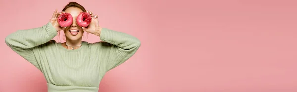 Mulher de camisola verde saindo da língua e segurando donuts perto do rosto isolado em rosa, banner — Fotografia de Stock
