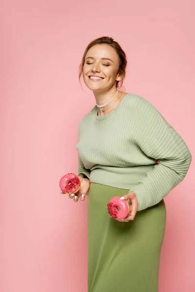 Sorridente donna incinta in possesso di gustose ciambelle isolate su rosa — Foto stock