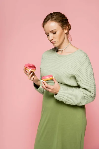 Donna incinta pensierosa che tiene ciambelle dolci isolate su rosa — Foto stock