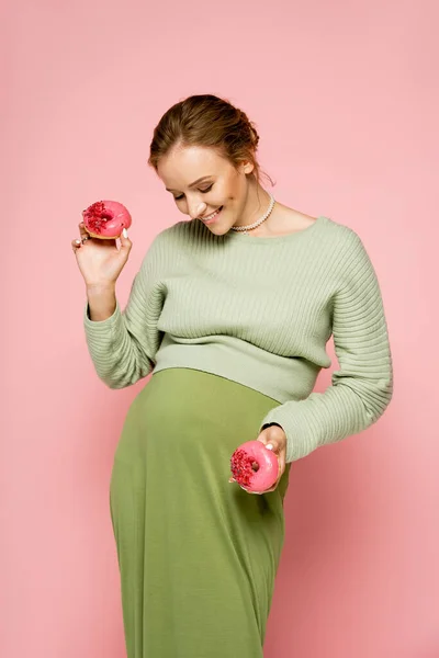 Mujer embarazada en elegante traje verde con rosquillas sobre fondo rosa - foto de stock