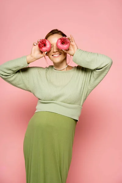 Positive schwangere Frau hält köstliche Donuts in Gesichtsnähe auf rosa Hintergrund — Stockfoto