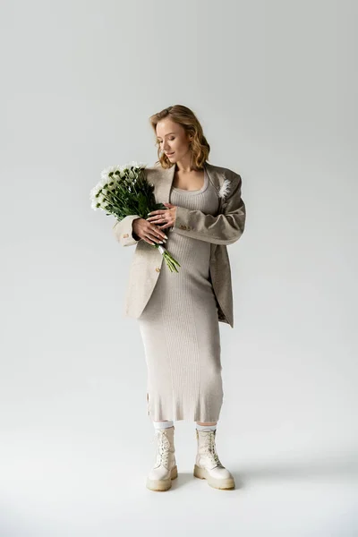 Pleine longueur de femme enceinte à la mode en robe et veste tenant des fleurs sur fond gris — Photo de stock