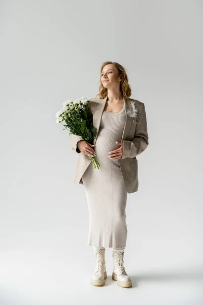 Femme enceinte à la mode en robe et veste tenant des fleurs blanches sur fond gris — Photo de stock