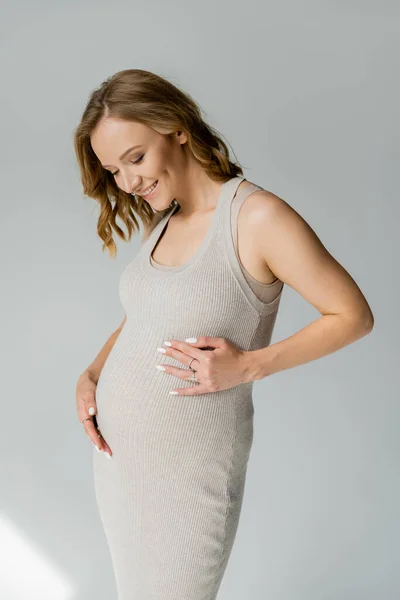 Mulher grávida positiva no vestido tocando barriga no fundo cinza — Fotografia de Stock