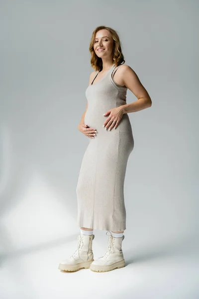 Повна довжина стильної вагітної жінки в одязі, дивлячись на камеру на сірому фоні — стокове фото