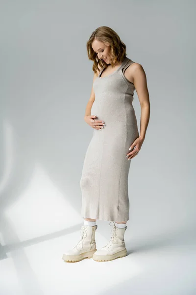 Pretty pregnant woman in beige dress and boots touching belly on grey background — Stock Photo