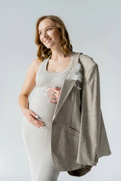 Модная беременная женщина в платье и пиджаке, касающаяся живота, изолированного на серой — стоковое фото