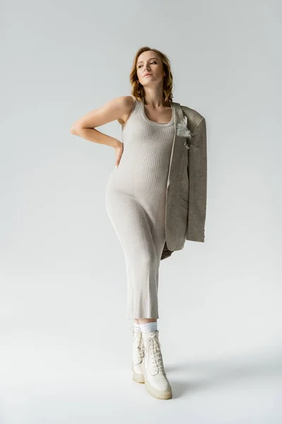 Модная беременная женщина в платье и куртке позирует на сером фоне — стоковое фото