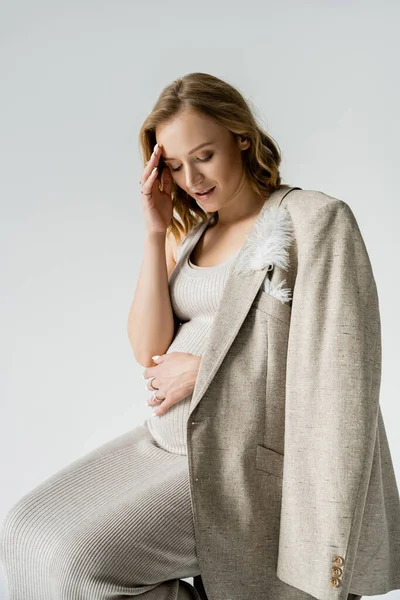 Молода вагітна жінка в сукні і піджаку дивиться на живіт ізольовано на сірому — стокове фото