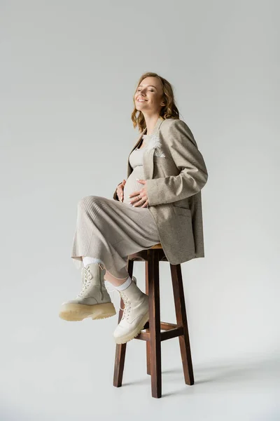 Mulher grávida agradável em vestido elegante sentado na cadeira em fundo cinza — Fotografia de Stock