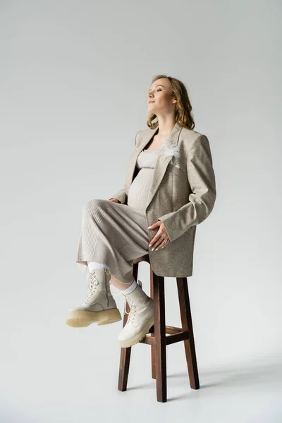 Jovem grávida de vestido e jaqueta sentada na cadeira em fundo cinza — Fotografia de Stock