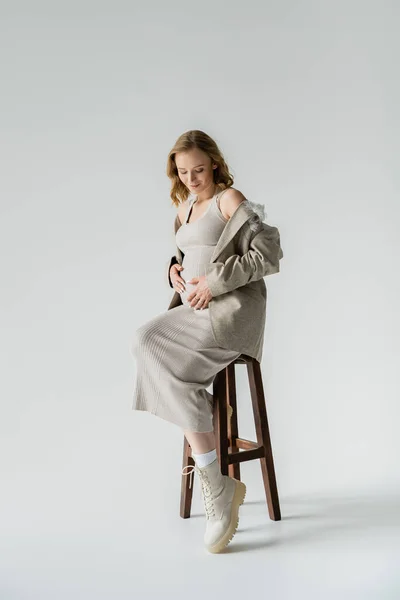 Longitud completa de la mujer embarazada positiva en el vestido sentado en la silla sobre fondo gris - foto de stock