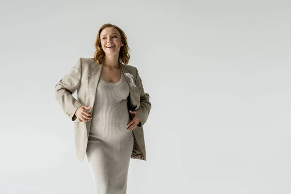 Happy pregnant woman in dress and jacket standing isolated on grey — Stock Photo