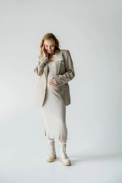 Full length of trendy pregnant woman in dress and boots standing on grey background — Stock Photo