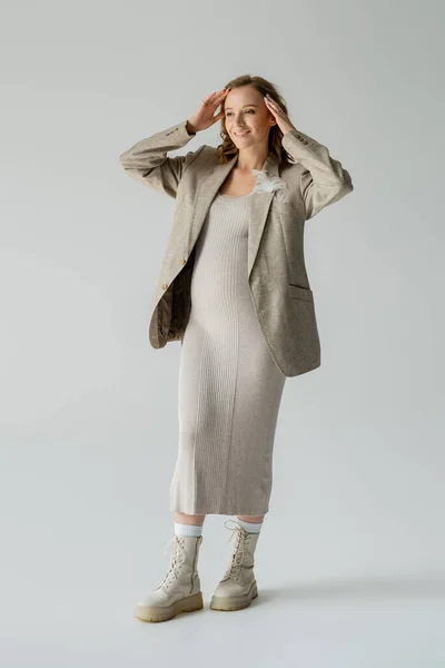 Volle Länge der fröhlichen schwangeren Frau in Kleid und Jacke berühren Haare auf grauem Hintergrund — Stockfoto
