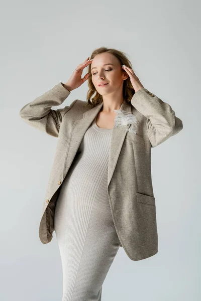 Stylish pregnant woman in beige dress and jacket with feather touching hair isolated on grey — Stock Photo