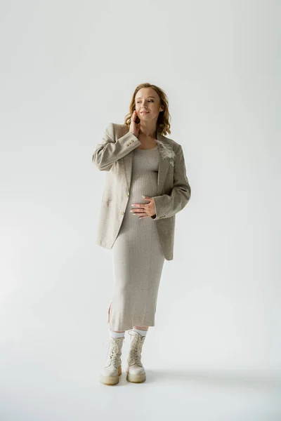 Longitud completa de la mujer embarazada de moda en botas y chaqueta posando sobre fondo gris - foto de stock