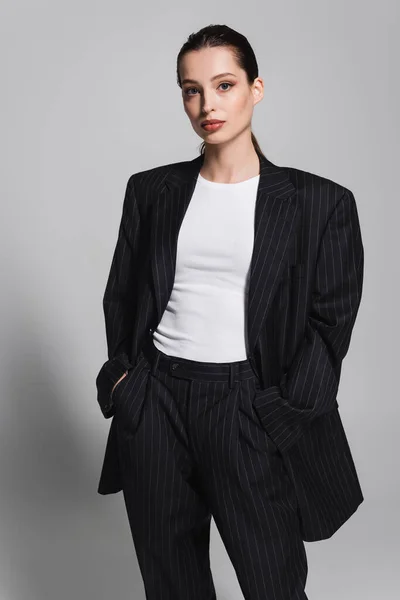 Trendige Frau im schwarzen Anzug posiert mit Händen in Taschen auf grauem Hintergrund — Stockfoto