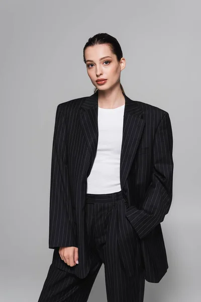 Trendy junge Frau im schwarzen Anzug posiert mit der Hand in der Tasche isoliert auf grau — Stockfoto