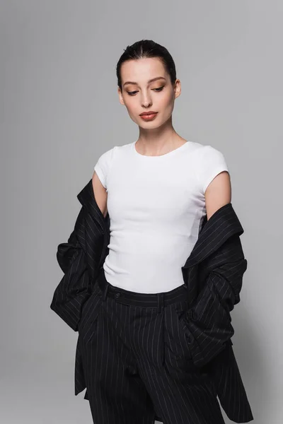 Donna di tendenza in t-shirt e completo nero che tiene le mani in tasche su sfondo grigio — Foto stock
