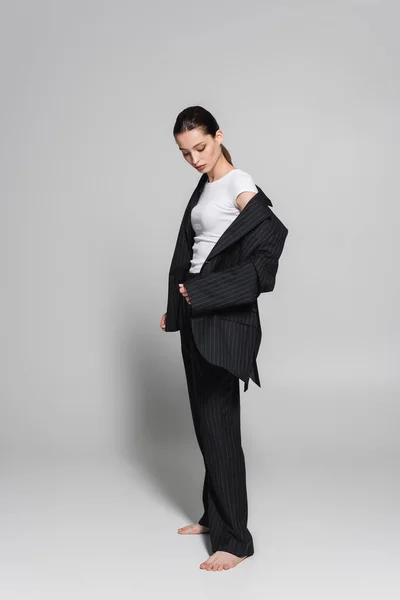 Longitud completa del modelo de moda en traje y camiseta mirando la manga de la chaqueta sobre fondo gris - foto de stock
