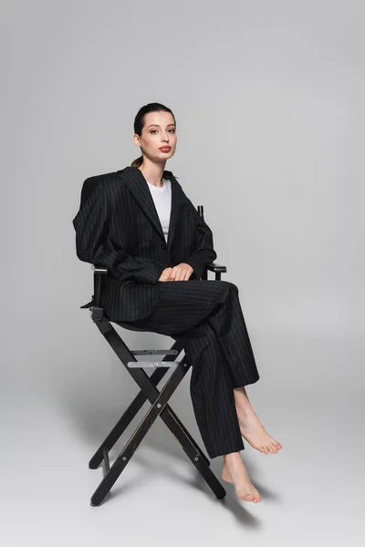 Longitud completa de mujer descalza en traje a rayas sentada en silla plegable sobre fondo gris - foto de stock
