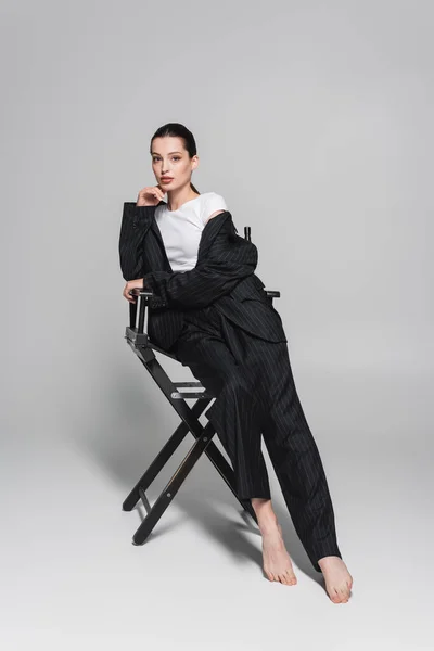 Barfuß-Model in gestreiftem Anzug und T-Shirt sitzt auf Klappstuhl auf grauem Hintergrund — Stockfoto