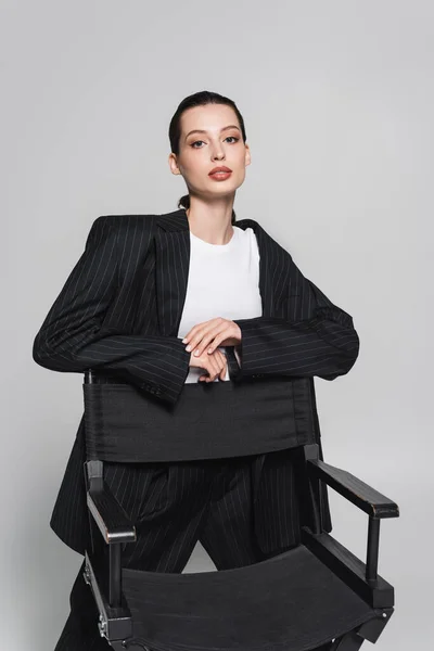 Гарненька брюнетка жінка стоїть біля розкладного крісла і дивиться на камеру ізольовано на сірому — стокове фото