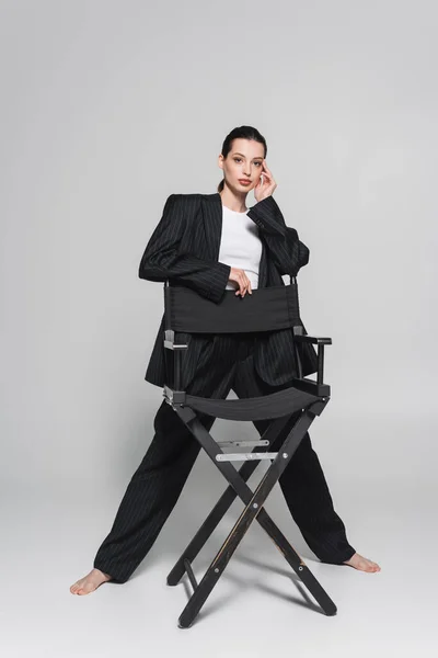 Pleine longueur de femme à la mode en costume posant près de chaise pliante sur fond gris — Photo de stock